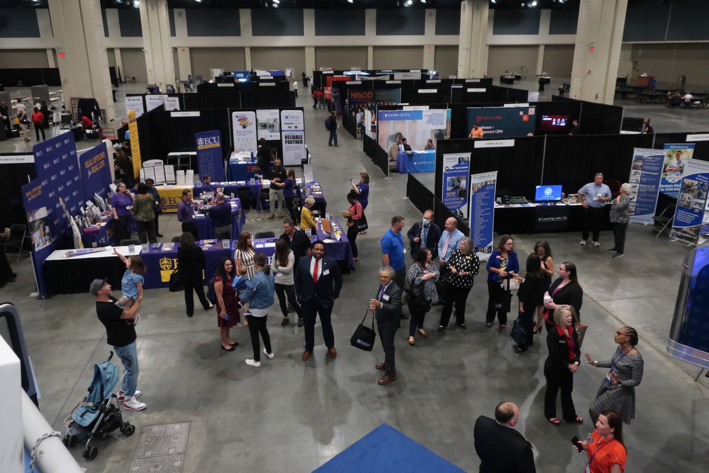 An overhead view of the conference floor, showing various information booths and several dozen attendees