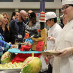 Several student chefs work an information table, demonstrating their food preparation for participants